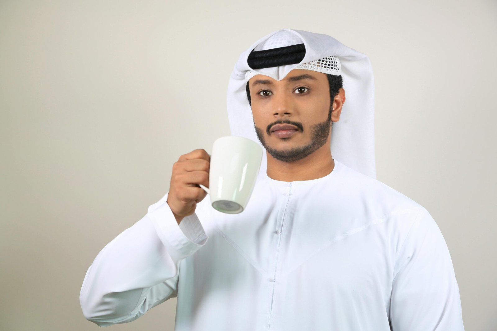 Young Middle Eastern man wearing traditional attire enjoys a cup of coffee indoors.